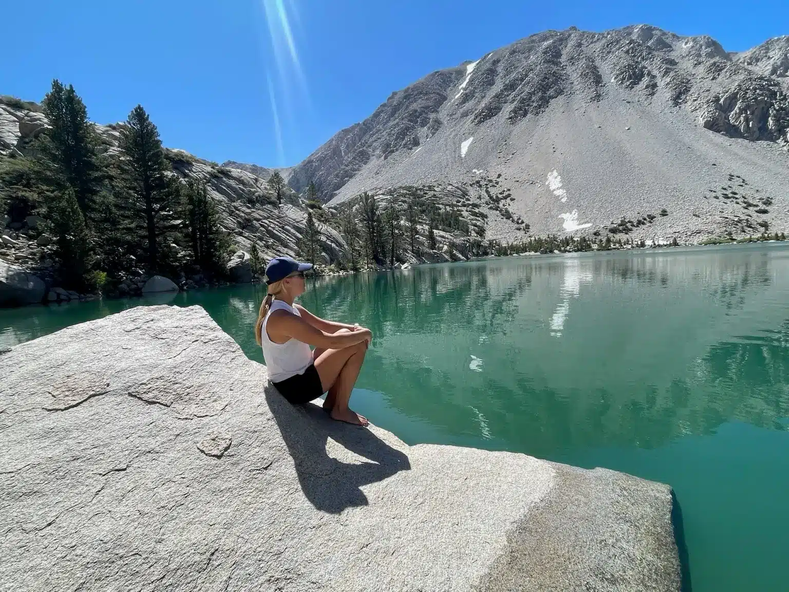 A person sitting on a rock by a turquoise lake, surrounded by trees and mountains under a clear blue sky near Mammoth Lakes, California.