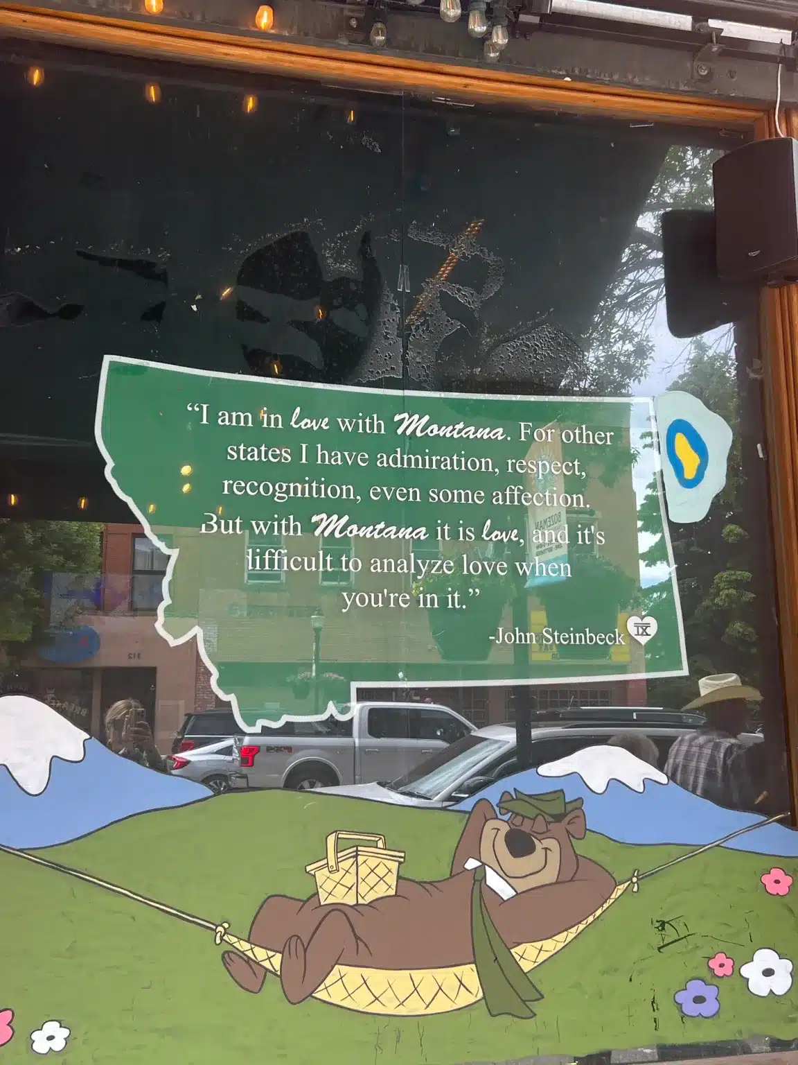 best-hotels-in-bozeman-montana-10 A window display in downtown Bozeman featuring a quote from John Steinbeck inside a green map of Montana: 'I am in love with Montana. For other states I have admiration, respect, recognition, even some affection. But with Montana it is love, and it's difficult to analyze love when you're in it.' Below the quote is an illustration of Yogi Bear relaxing in a hammock.