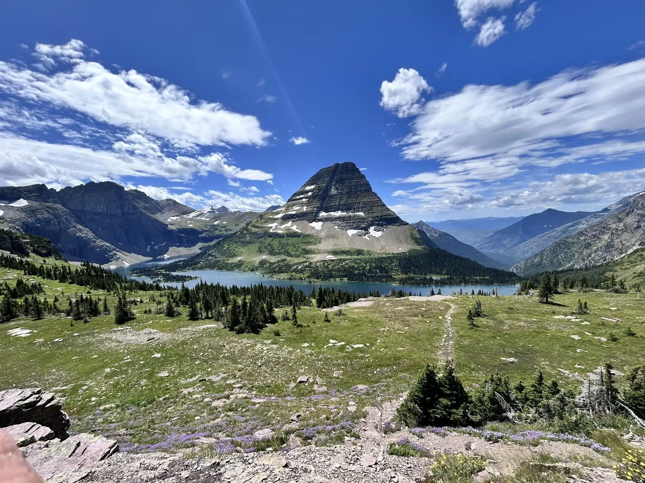 things-to-do-in-glacier-national-park-1 Pointed, layered mountain peak overlooking a blue alpine lake and green meadows under a blue sky. This view is of the Hidden Lake Trail which is one of the best hikes in Glacier National Park.