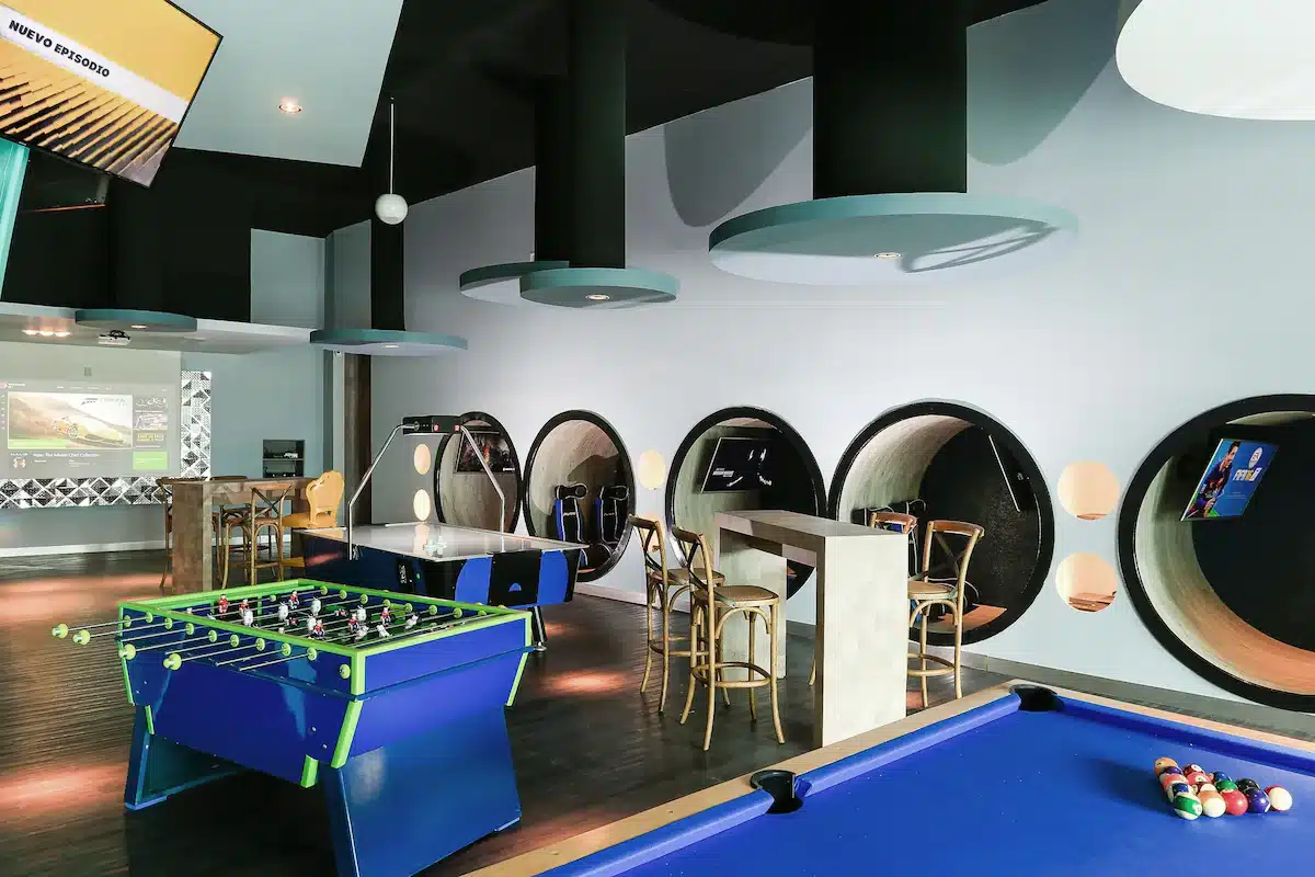 An interior view of a teen club room at an all-inclusive resort featuring a foosball table, air hockey table, and a billiard table. The room includes round window-like features and modern lighting.