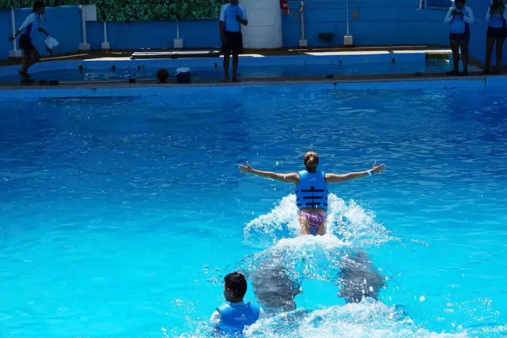 A person in a life jacket with arms outstretched is swimming in a blue pool, while another individual partially visible is in the water below them.