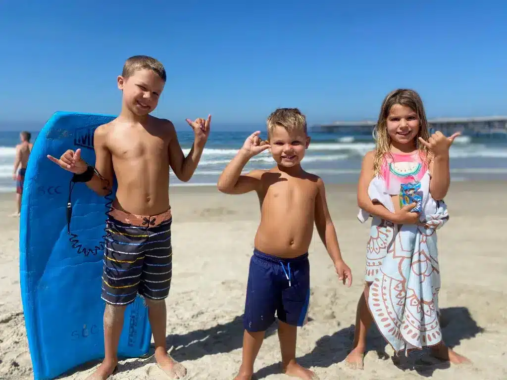 Three young kids standing on a beach with boogie boards during their family summer vacation