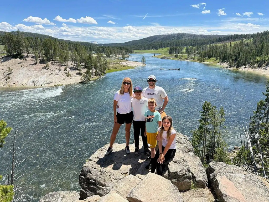 A family posing on a rock with a river and mountains behind them while on an affordable family vacation in Yellowstone
