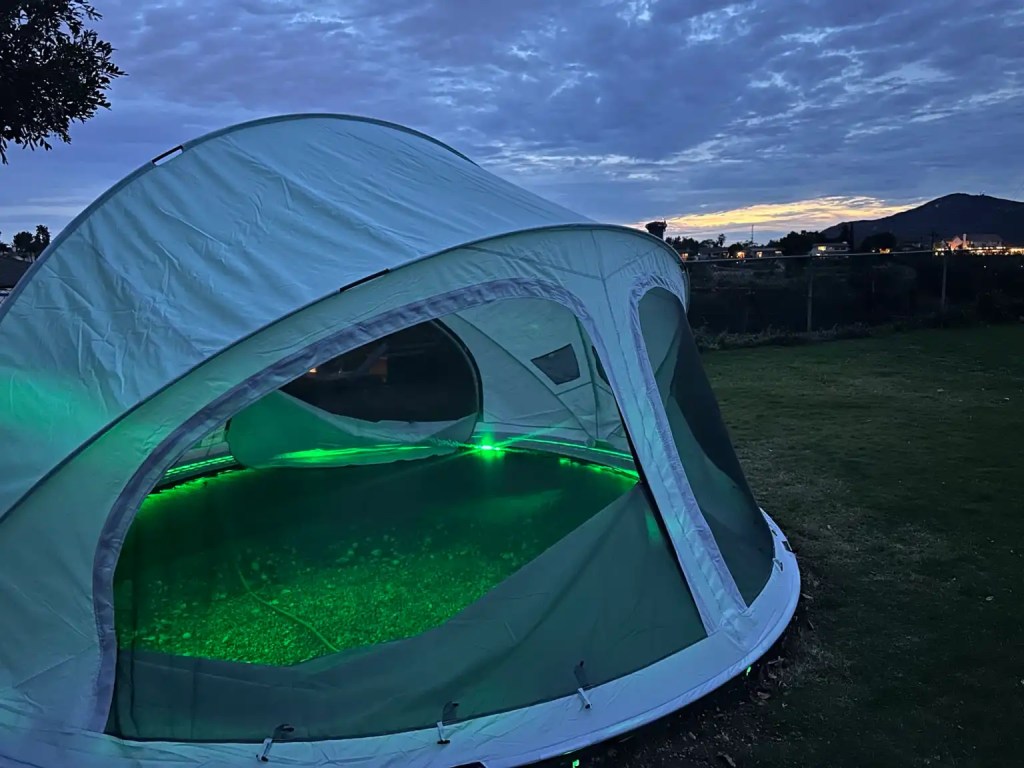 A green-lit dome tent set up in a backyard during twilight, with a clear view through its mesh walls.