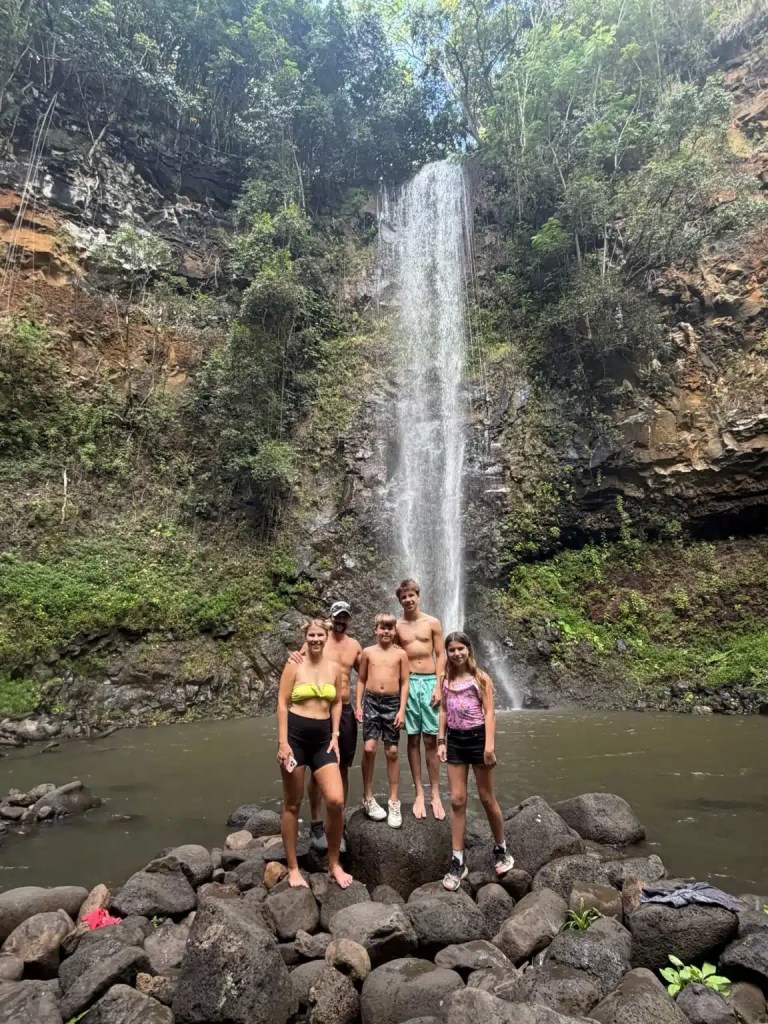 Group of five people standing on rocks in front of a waterfall in Kauai, surrounded by lush greenery.