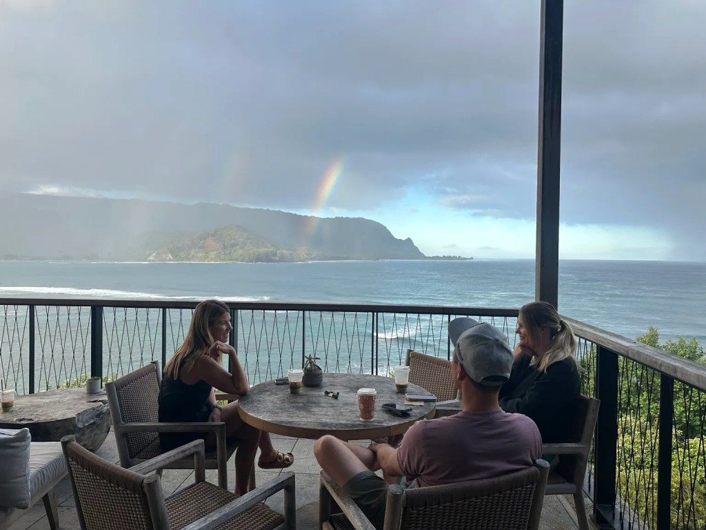Three people enjoying coffee on a balcony at 1 Hotel Hanalei Resort overlooking Hanalei Bay, with a rainbow visible in the background and lush mountains in sight.