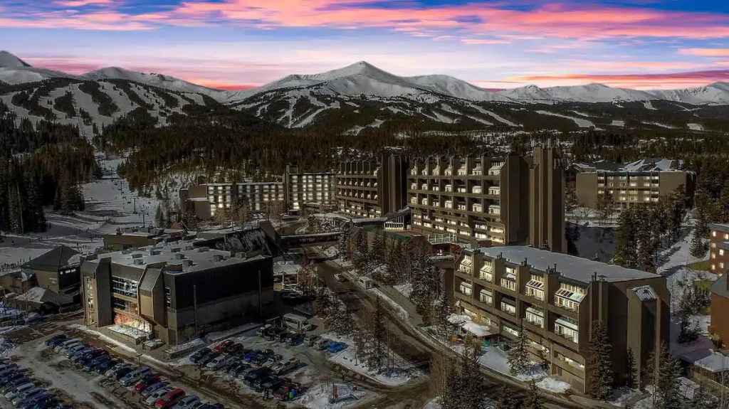 Aerial view of a hotel complex in Breckenridge, Colorado, surrounded by snow-covered mountains and trees under a colorful sunset sky.
