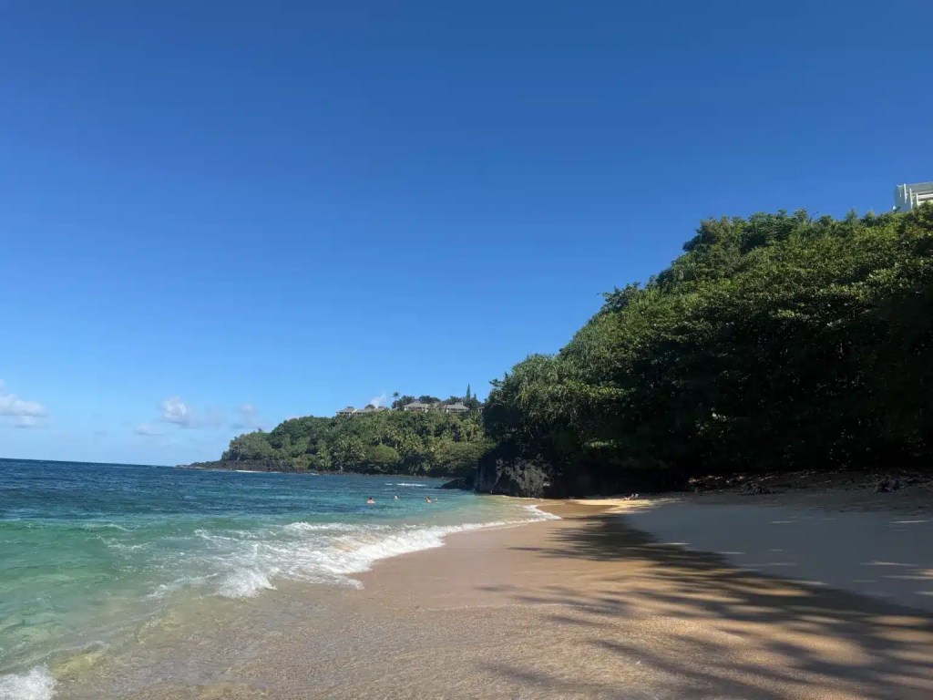 A scenic view of Hideaway Beach on Kauai with turquoise water, golden sand, lush green cliffs, and bright blue skies.