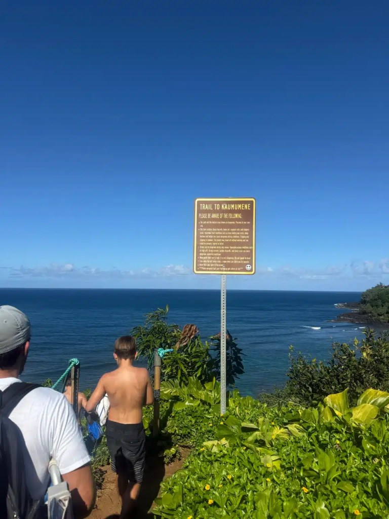 Two people approaching a trail sign that reads 'Trail to Kaumumene,' overlooking the ocean with a clear blue sky and lush greenery.