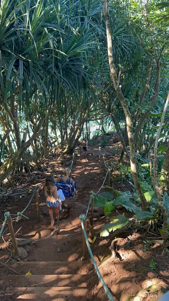 Two children walking down a narrow, uneven trail surrounded by lush greenery leading to Hideaway Beach in Kauai.