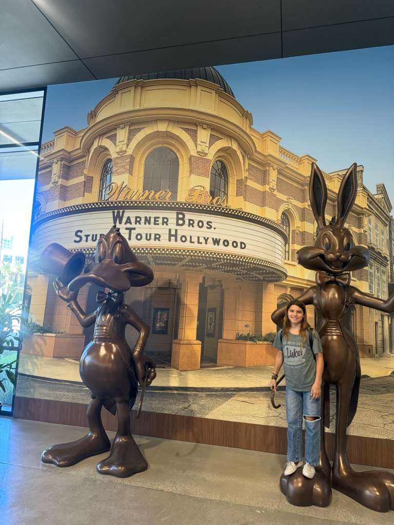 A young girl stands between two bronze statues of animated characters from Warner Bros., with a large promotional backdrop of the Warner Bros. Studio Tour Hollywood.