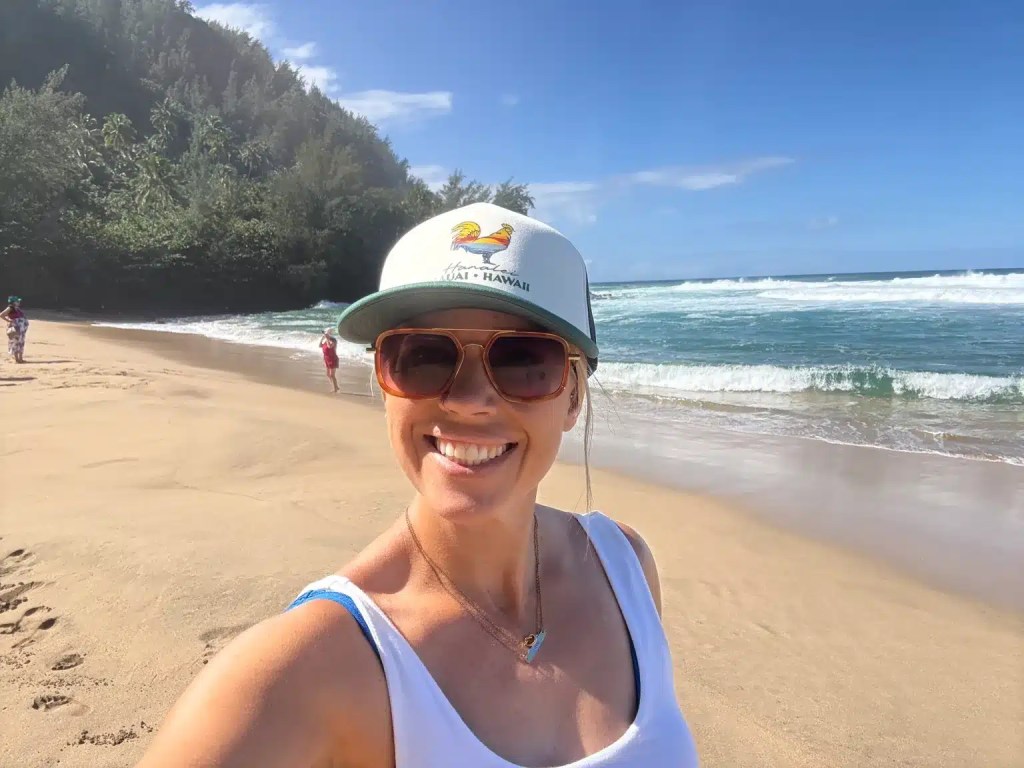 A person smiling on a sandy Kauai beach with ocean waves and lush greenery in the background.