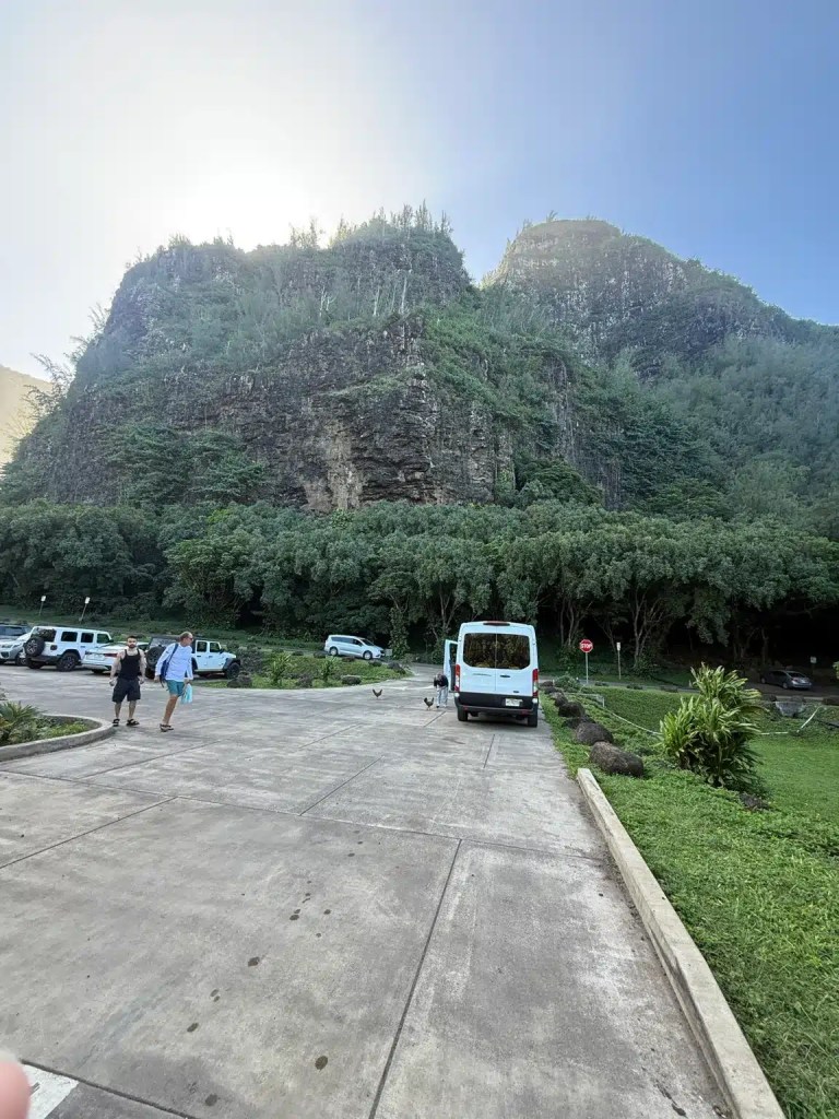 Parking area with vehicles and people walking towards the entrance of Ke‘e Beach. Lush green cliffs rise in the background under a clear sky.