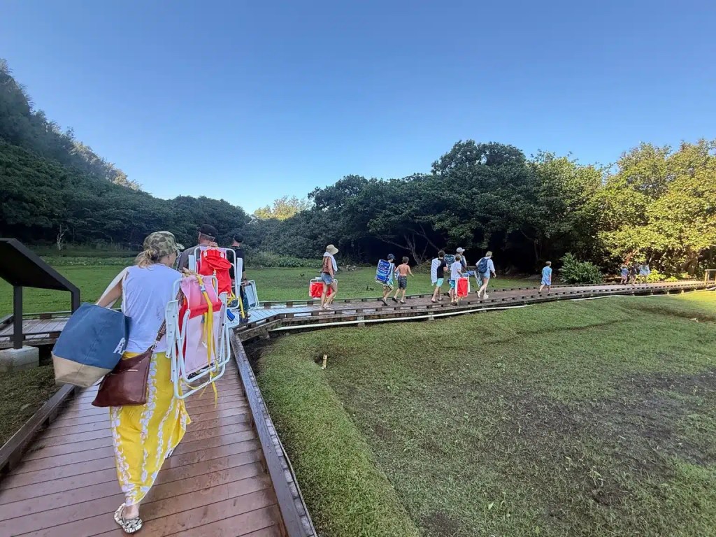 People carrying beach gear walking along a wooden path towards Ke‘e Beach in Kauai, surrounded by lush greenery under a clear blue sky.