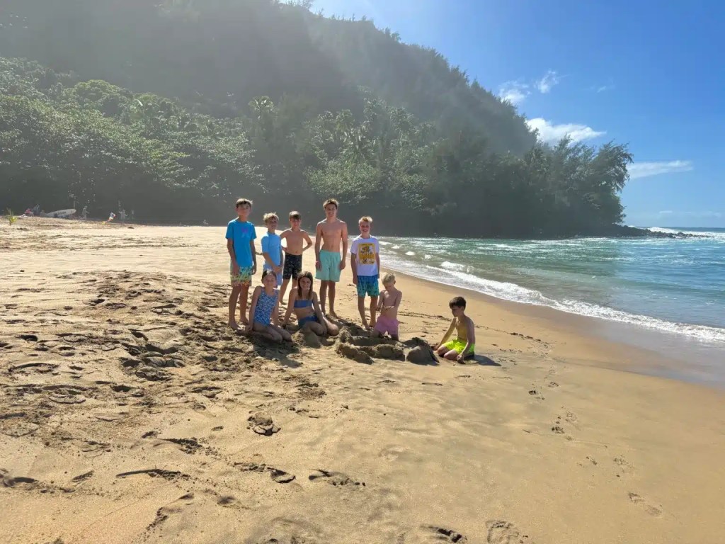 A group of children gathered on the sandy beach in Kauai, enjoying a sunny day by the ocean, with lush greenery and waves in the background.