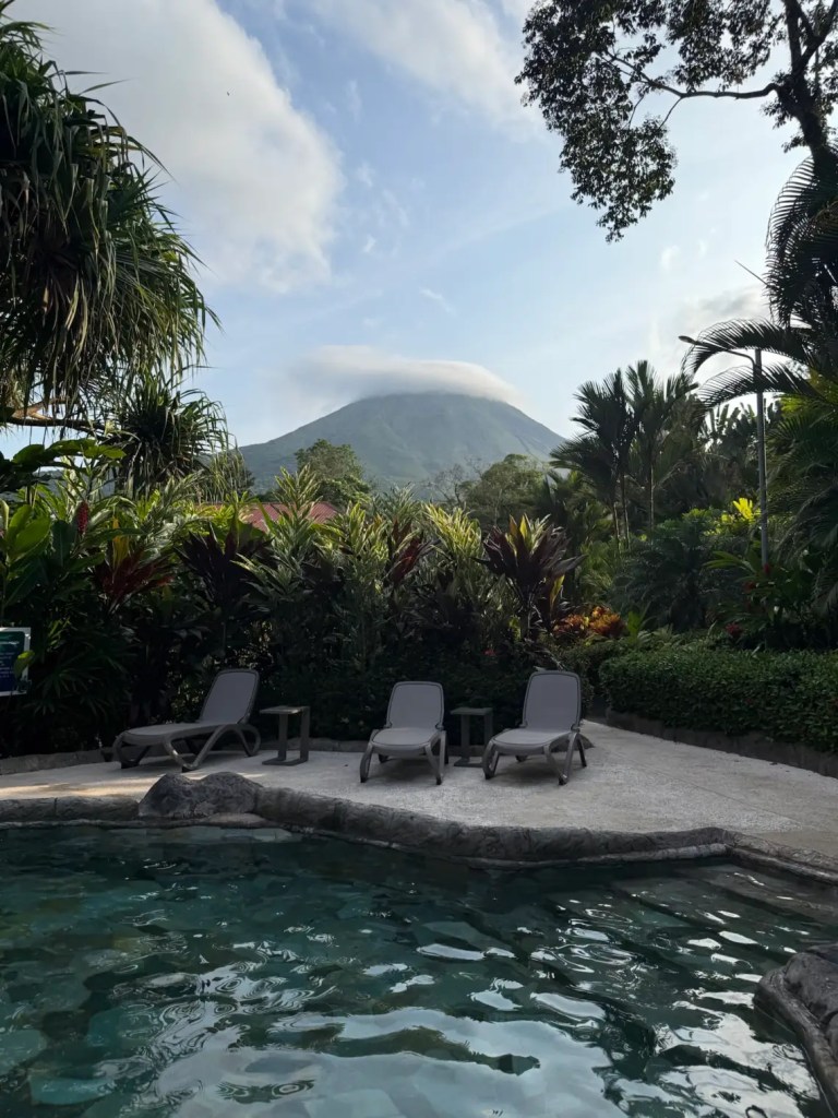 A scenic view of Arenal Volcano in the background with a pool area featuring lounge chairs and lush tropical plants in the foreground.