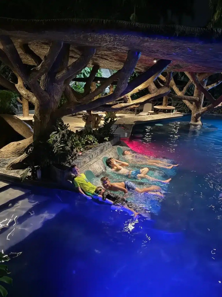 Children relaxing in a Costa Rica hot spring at night, surrounded by vibrant lights and a tree canopy.