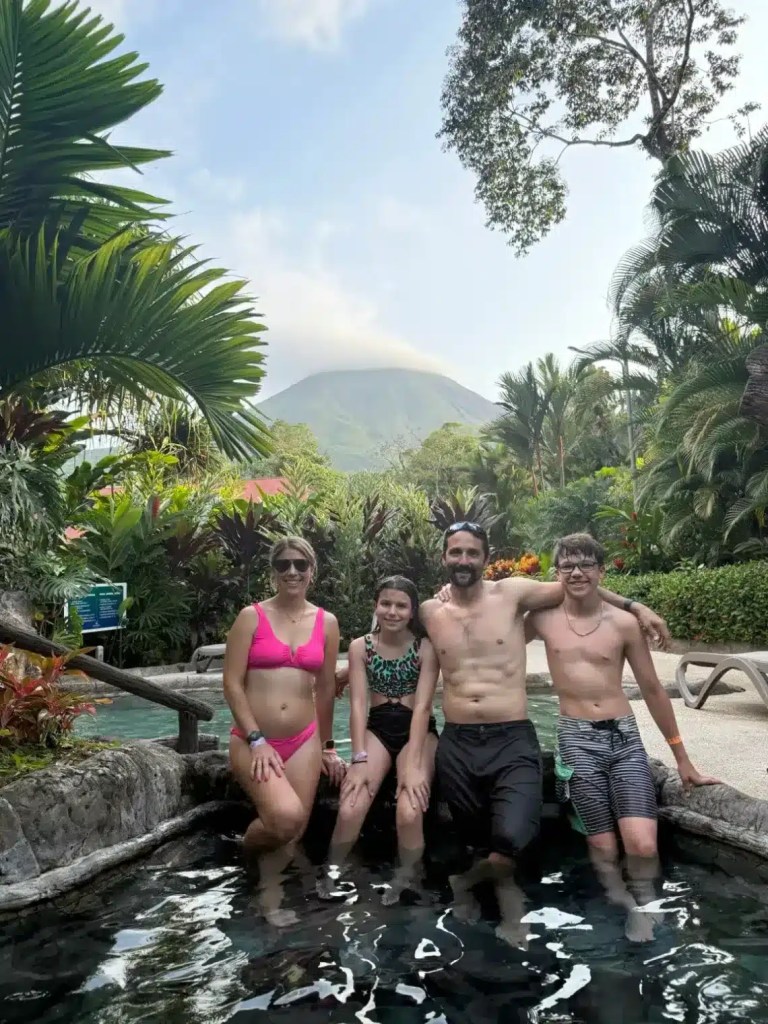 Four people enjoying a hot spring in La Fortuna, Costa Rica, with Arenal Volcano in the background surrounded by lush greenery.