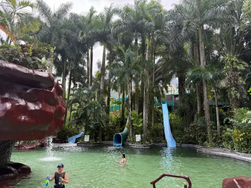A tropical hot springs resort in La Fortuna, Costa Rica, featuring palm trees, swimming areas, and water slides, with guests enjoying the warm water.