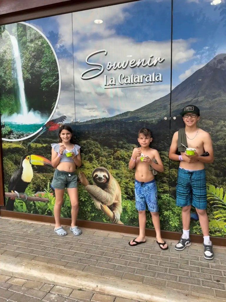 Three children stand in front of a souvenir shop with a vibrant mural featuring a waterfall, a toucan, and a sloth, showcasing the natural beauty of La Fortuna, Costa Rica.