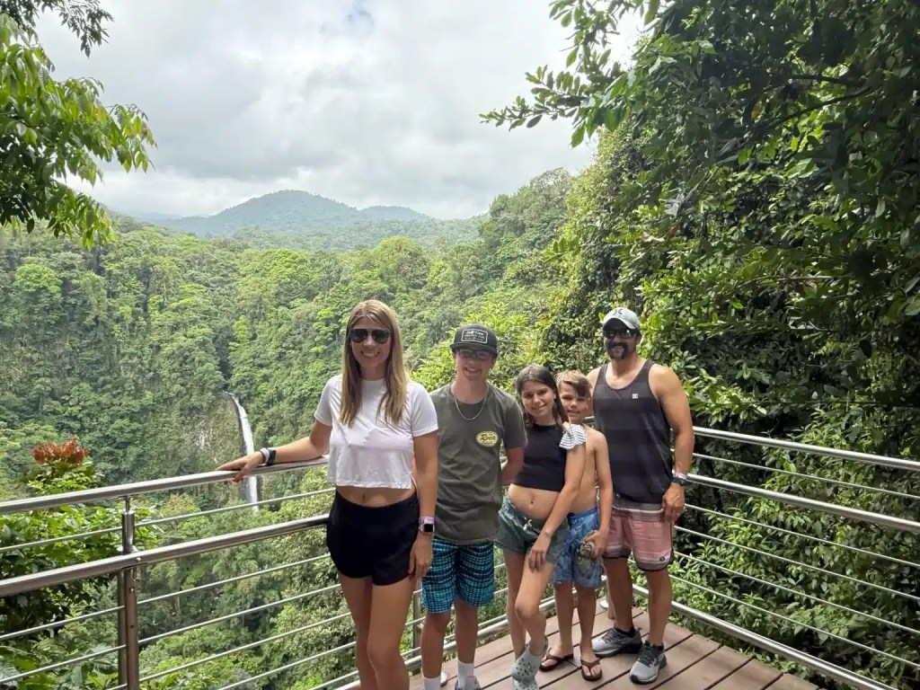 A family stands on a viewing platform at La Fortuna, Costa Rica, with a lush green landscape and a waterfall in the background.