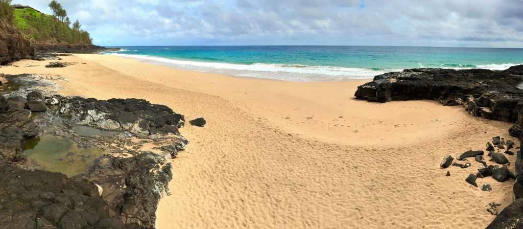 A tranquil view of Secret Beach in Kauai, showcasing golden sand, rocky cliffs, and the blue Pacific Ocean under partly cloudy skies.