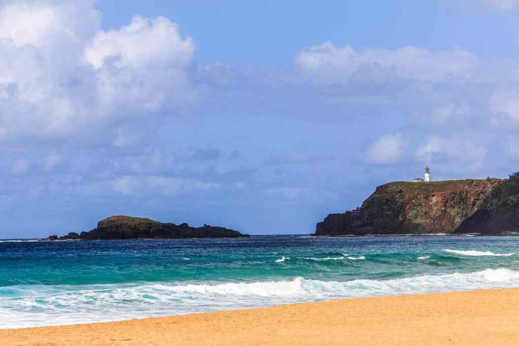 A scenic view of Secret Beach in Kauai, featuring golden sand, turquoise waves, and a lighthouse perched on a green cliff in the distance under a partly cloudy sky.