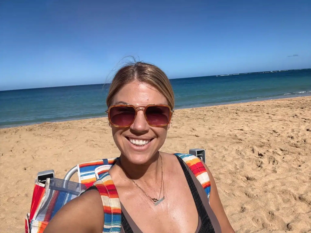 A person smiling on a sandy Kauai beach with the ocean in the background, wearing sunglasses and a swimsuit.