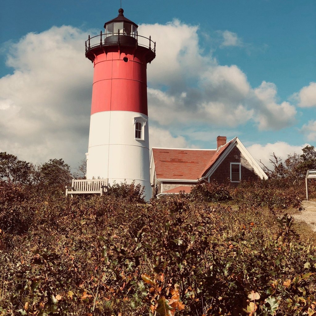 A red and white striped lighthouse stands next to a coastal house in Cape Cod, surrounded by lush greenery and under a blue sky with scattered clouds.