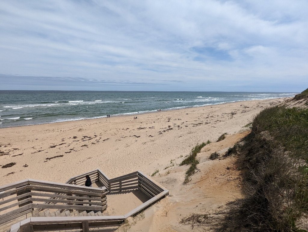 A sandy beach in Cape Cod, featuring wooden stairs leading down to the shoreline, with gentle waves lapping at the shore under a cloudy sky.