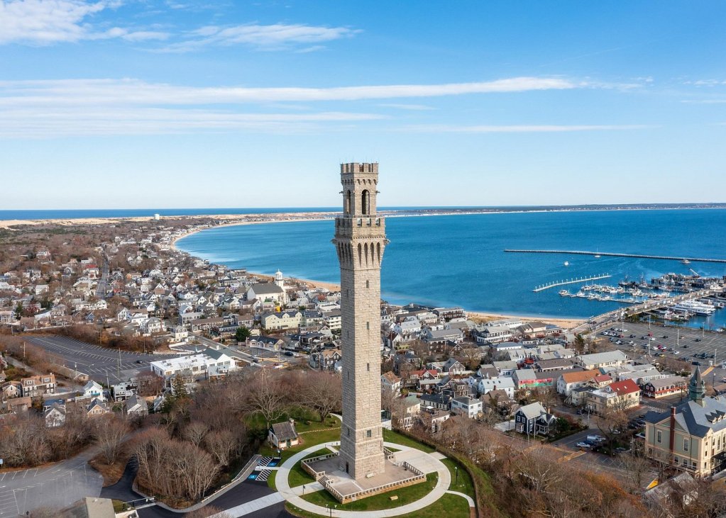 Aerial view of the Pilgrim Monument in Provincetown, Massachusetts, overlooking the coast and harbor. Scenic backdrop featuring blue water and sandy beach with surrounding town homes.