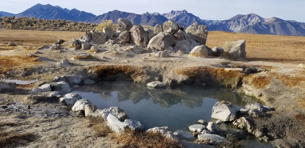 A natural hot spring surrounded by rocks and dry grass, with mountains in the background.