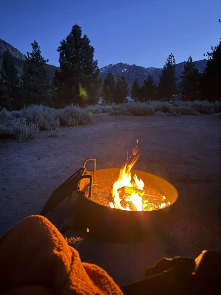 A cozy campfire burning in a fire pit at dusk, surrounded by trees and mountains in Mammoth Lakes, California.