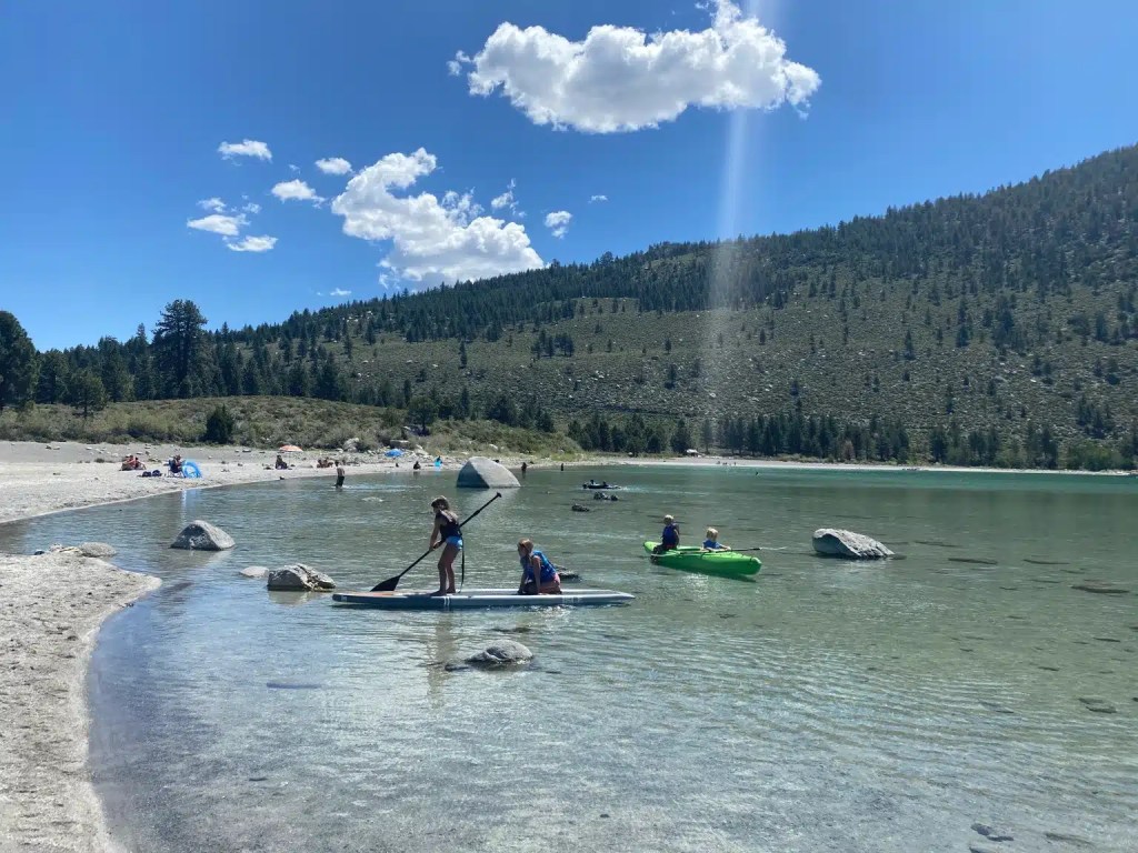 People paddleboarding and kayaking on a clear lake surrounded by greenery, with some beachgoers relaxing on the shore.