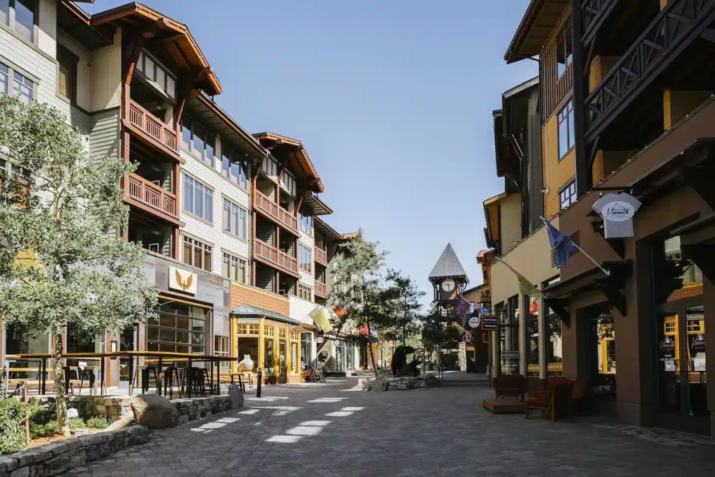 A pedestrian-friendly street in Mammoth Mountain Village, showcasing colorful buildings, outdoor seating, and tree-lined paths under a clear blue sky.