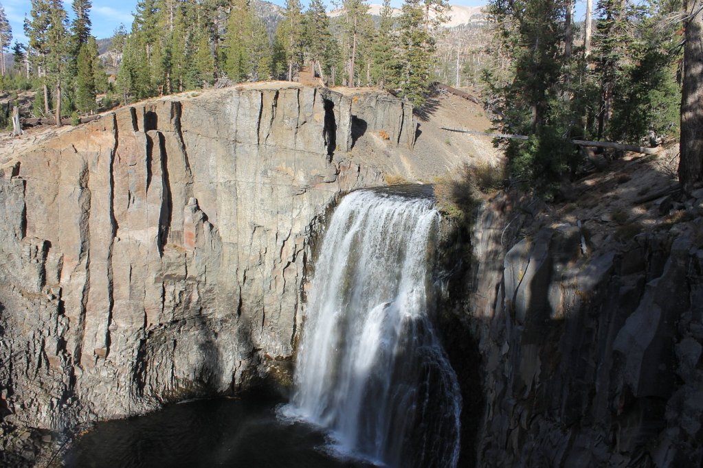 A scenic view of Rainbow Falls, showcasing cascading water surrounded by rocky cliffs and pine trees in a picturesque natural setting.