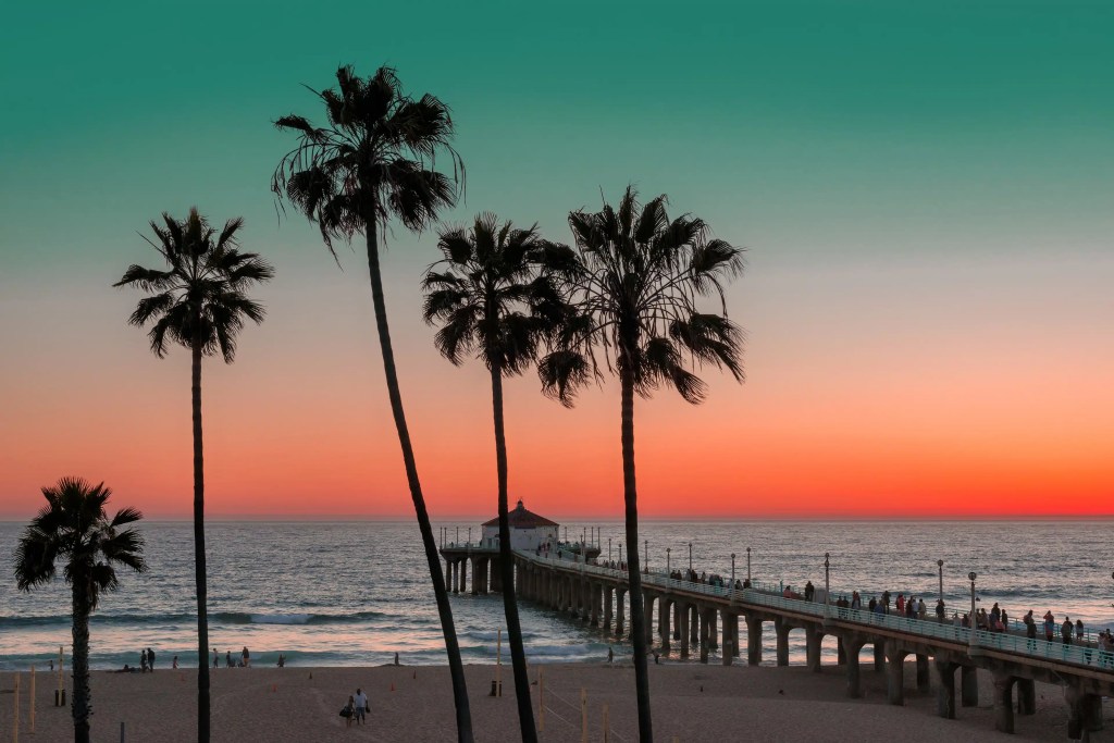 A scenic view of a beach pier at sunset, with silhouetted palm trees in the foreground and a colorful sky transitioning from blue to pink and orange.