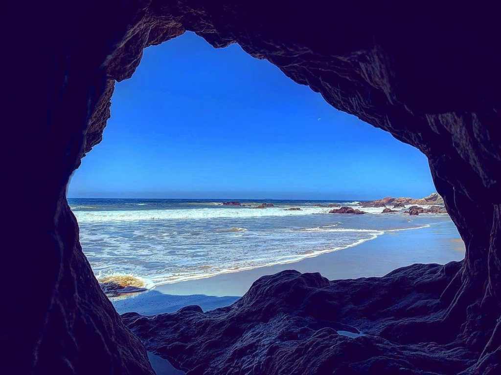 View of the ocean and a sandy Los Angeles beach through a rocky cave with waves crashing in the background and a clear blue sky.