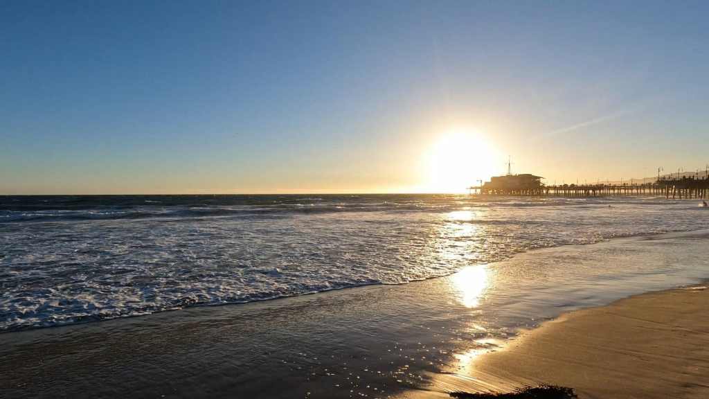 Sunset over a Los Angeles beach with gentle waves lapping at the shore, and a pier in the distance.
