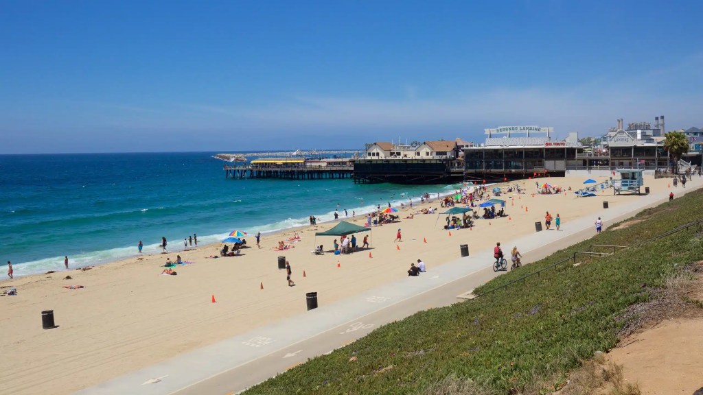 A sunny beach scene at Redondo Beach, with people enjoying the sand and ocean. There are colorful umbrellas and beachgoers near the pier, which features shops and restaurants in the background.