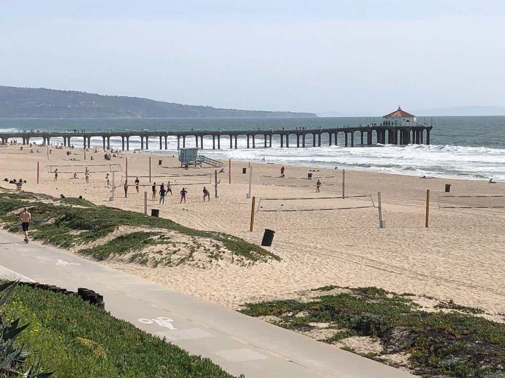 A view of a wide sandy beach with volleyball courts and people enjoying the sun, alongside a wooden pier with a pagoda-style roof.