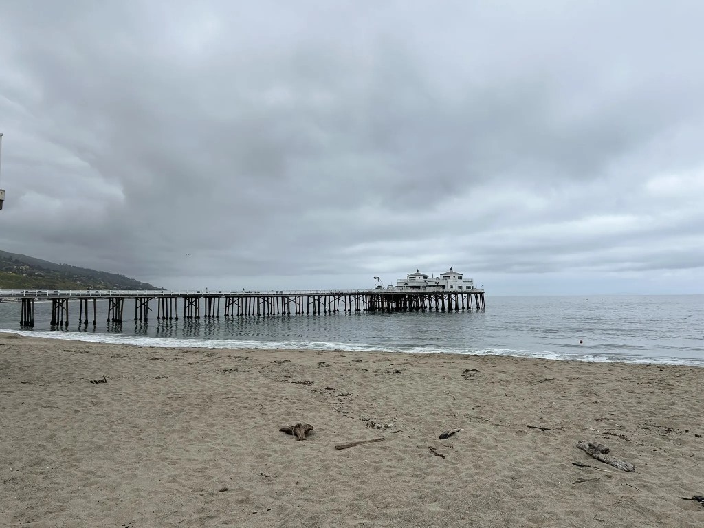 A sandy Los Angeles beach with a wooden pier extending into the ocean under a cloudy sky.