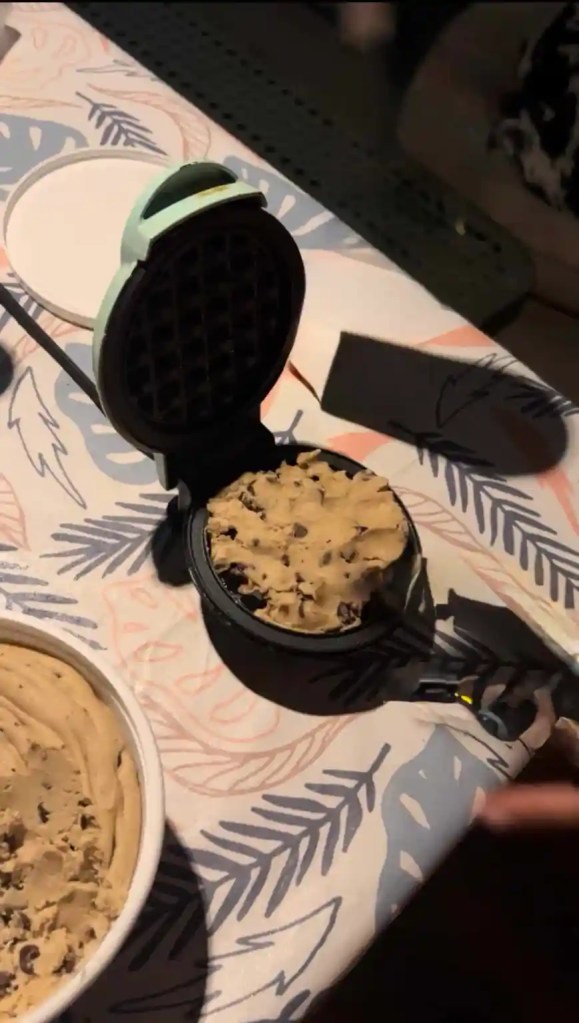 A waffle maker with cookie dough inside, positioned on a patterned tablecloth, with a bowl of cookie dough nearby.