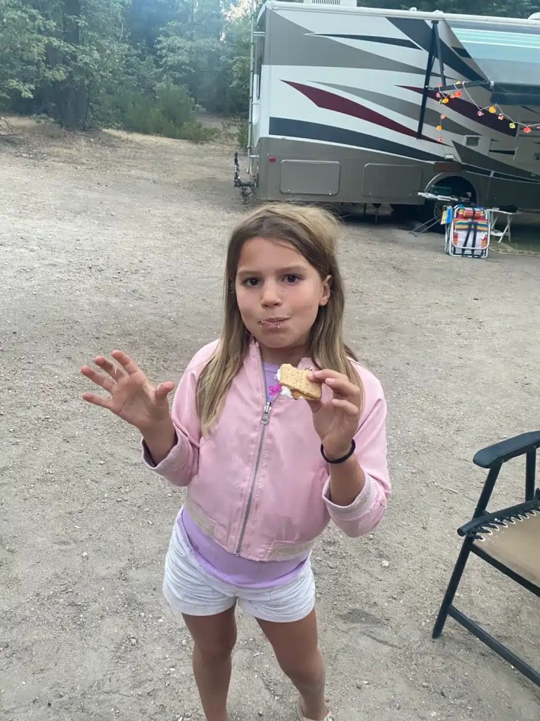 A young girl wearing a pink jacket and white shorts is standing outdoors at a campsite, holding an easy camping dessert and showing a playful expression.