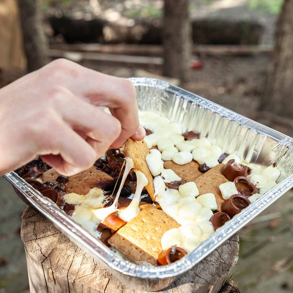 A hand reaching into a foil pan filled with an easy camping dessert of melted chocolate, marshmallows, and graham crackers, with gooey marshmallow stretching from a cracker.