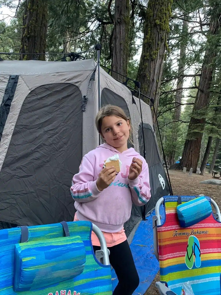 A young girl in a pink sweatshirt is smiling while holding an easy camping dessert, standing near a camping tent surrounded by trees. Two colorful beach chairs are visible in the foreground.