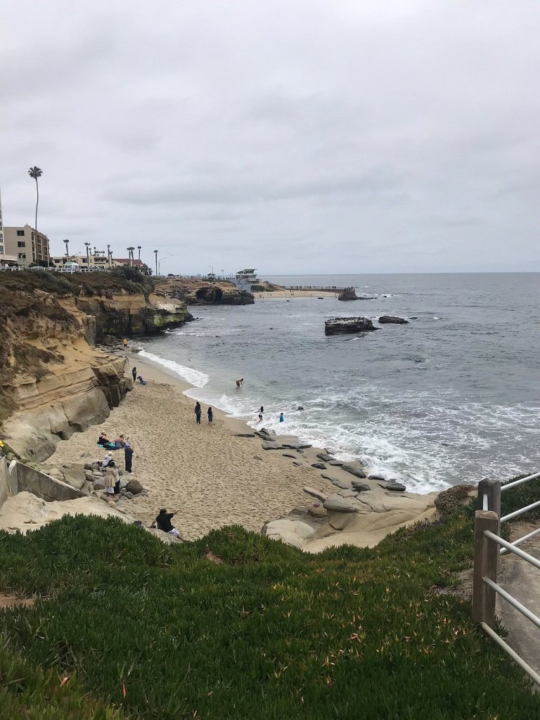 View of La Jolla Cove with sandy beach, rocky shoreline, and people enjoying the water under a cloudy sky.