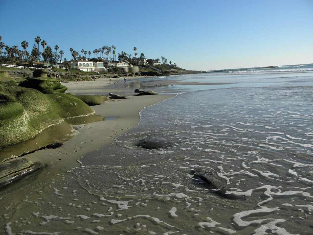 A tranquil beach scene at La Jolla with gentle waves lapping the shore, tide pools visible among the rocks, and a clear blue sky overhead.