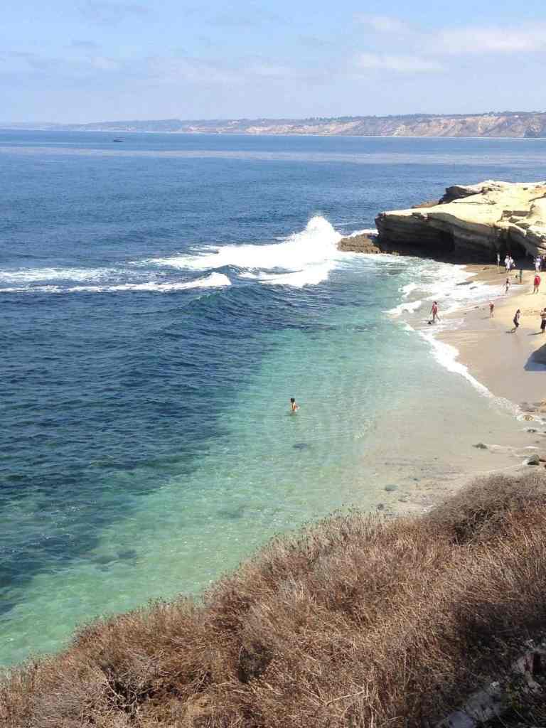 A scenic view of one of the La Jolla beaches in San Diego with clear blue water, gentle waves, and a few people enjoying the shoreline.