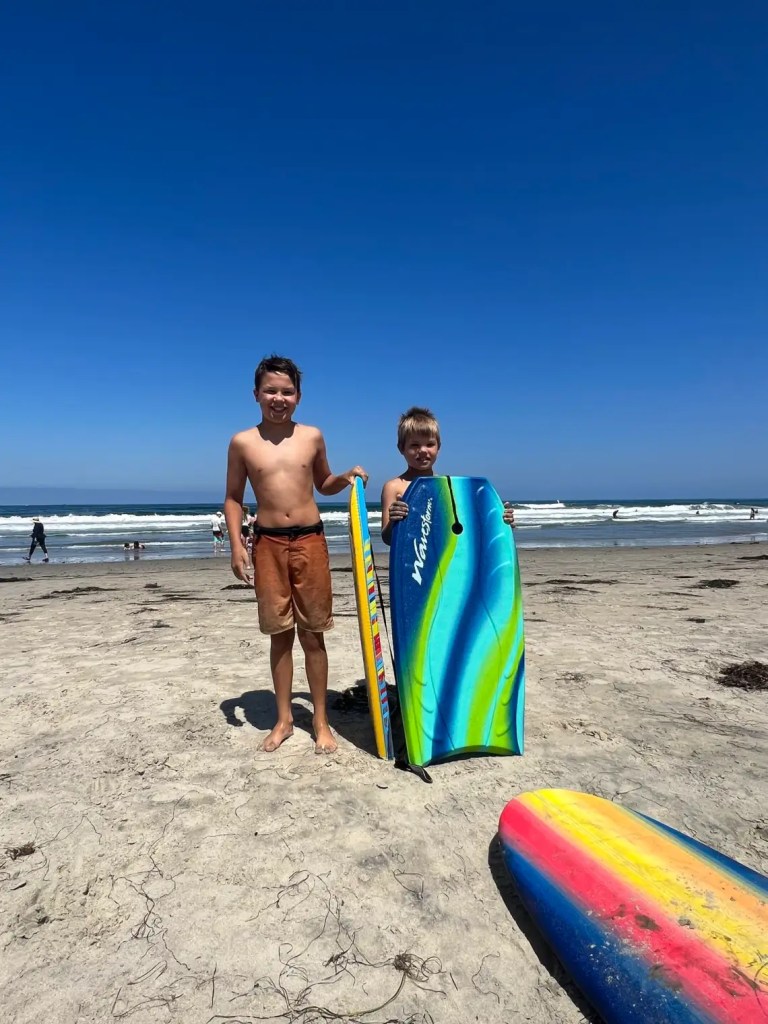 Two children standing on a sandy beach, holding colorful boogie boards, with the ocean and waves in the background and a clear blue sky above.